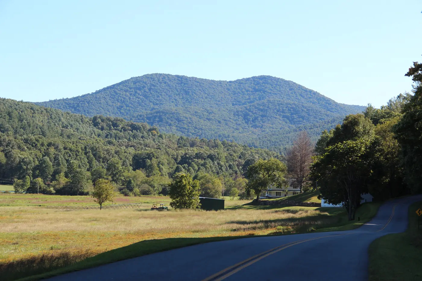 Rounded forested mountain above an open meadow and a curving rural road