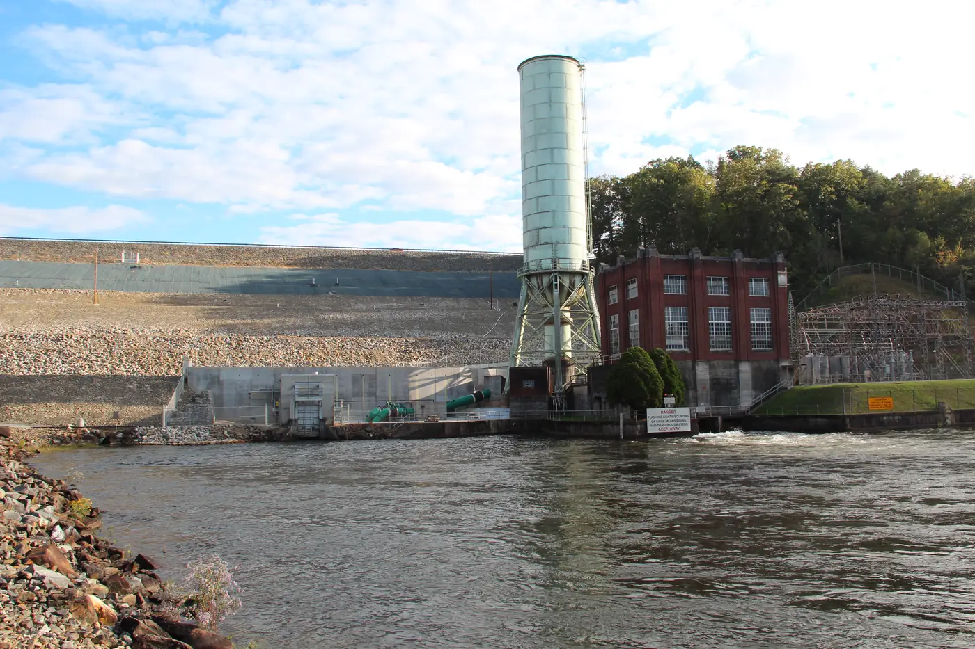 Rock-faced dam with a tall cylindrical surge tank and brick powerhouse beside the river