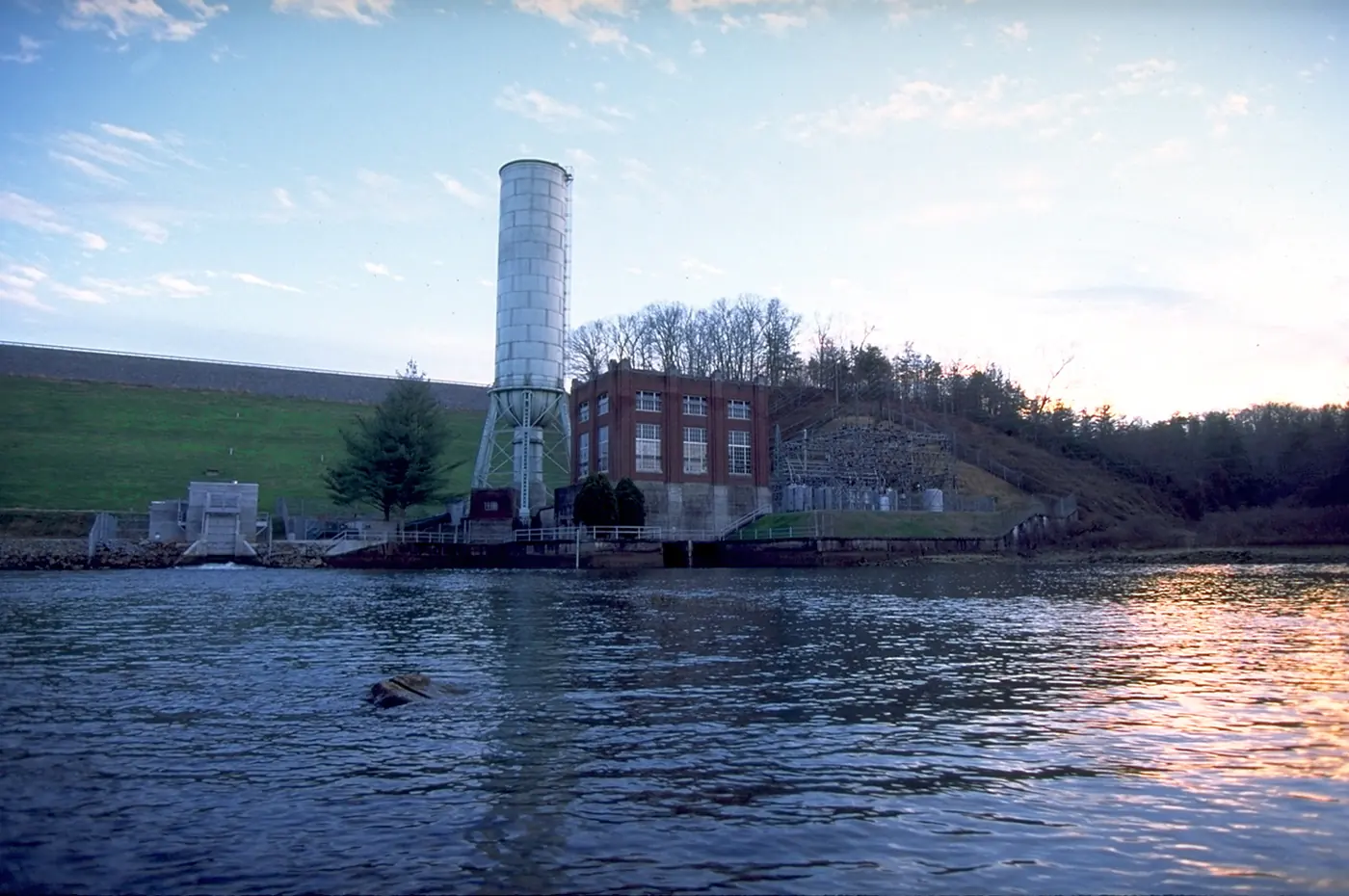 Grassy dam face, cylindrical surge tank and brick powerhouse reflected in the river