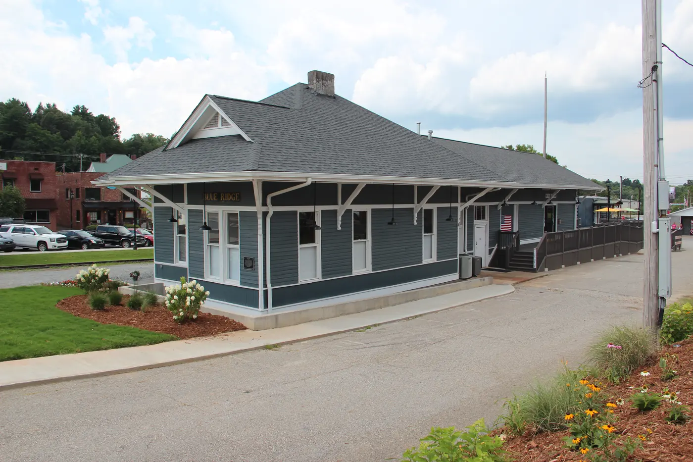 Gray-blue wooden depot building with white trim beside a paved lot and rail line