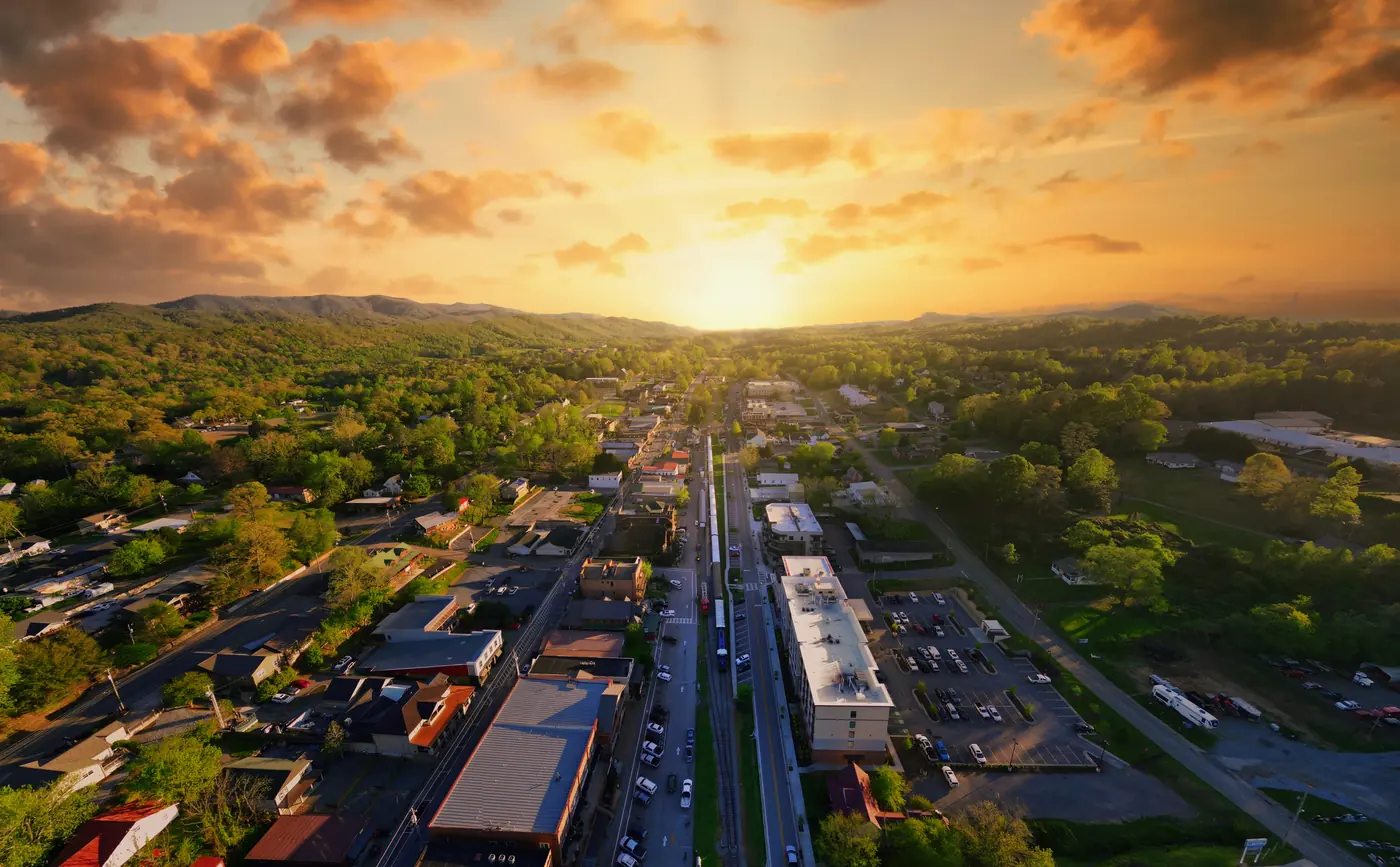Aerial view of a small mountain town main street and rail line at sunset, with wooded hills beyond
