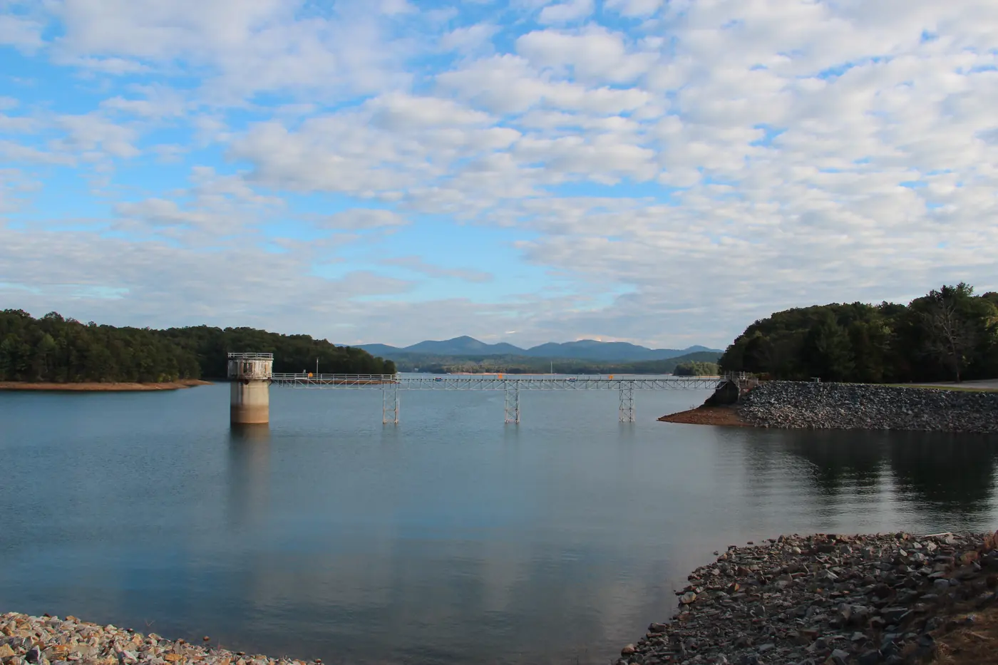 Calm lake with a concrete intake tower, wooded points and distant blue mountains under scattered clouds