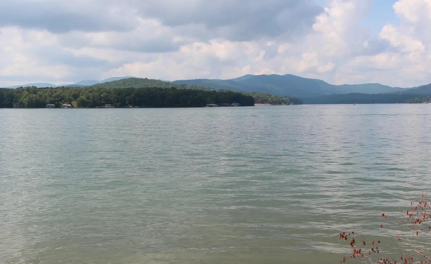Wide open lake water leading to a forested shoreline and layered blue mountains under white clouds