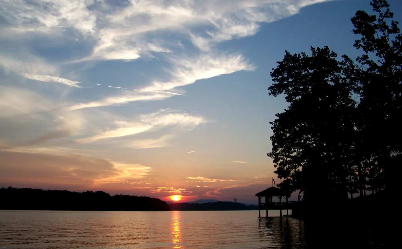 Sun setting over a lake behind a far ridge, with silhouetted trees and a gazebo on a dock at right