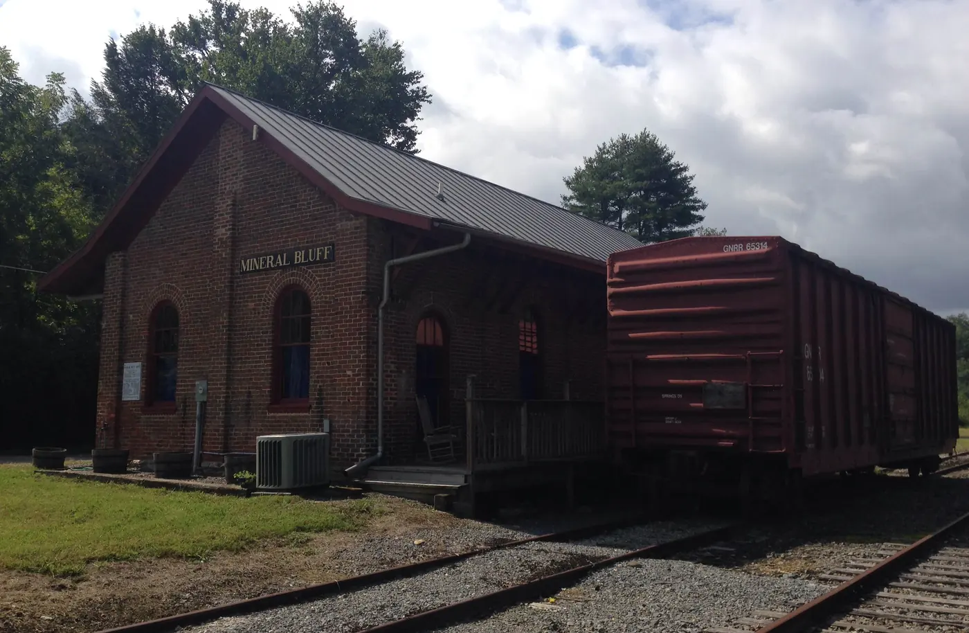Brick railroad depot with a metal roof beside a red boxcar on the tracks
