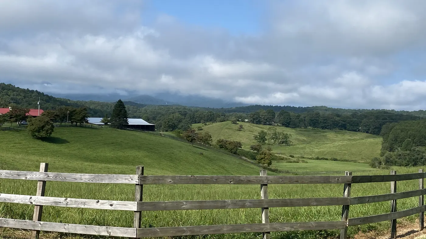 Rolling green pastures with a wooden fence, scattered barns and distant cloud-capped mountains