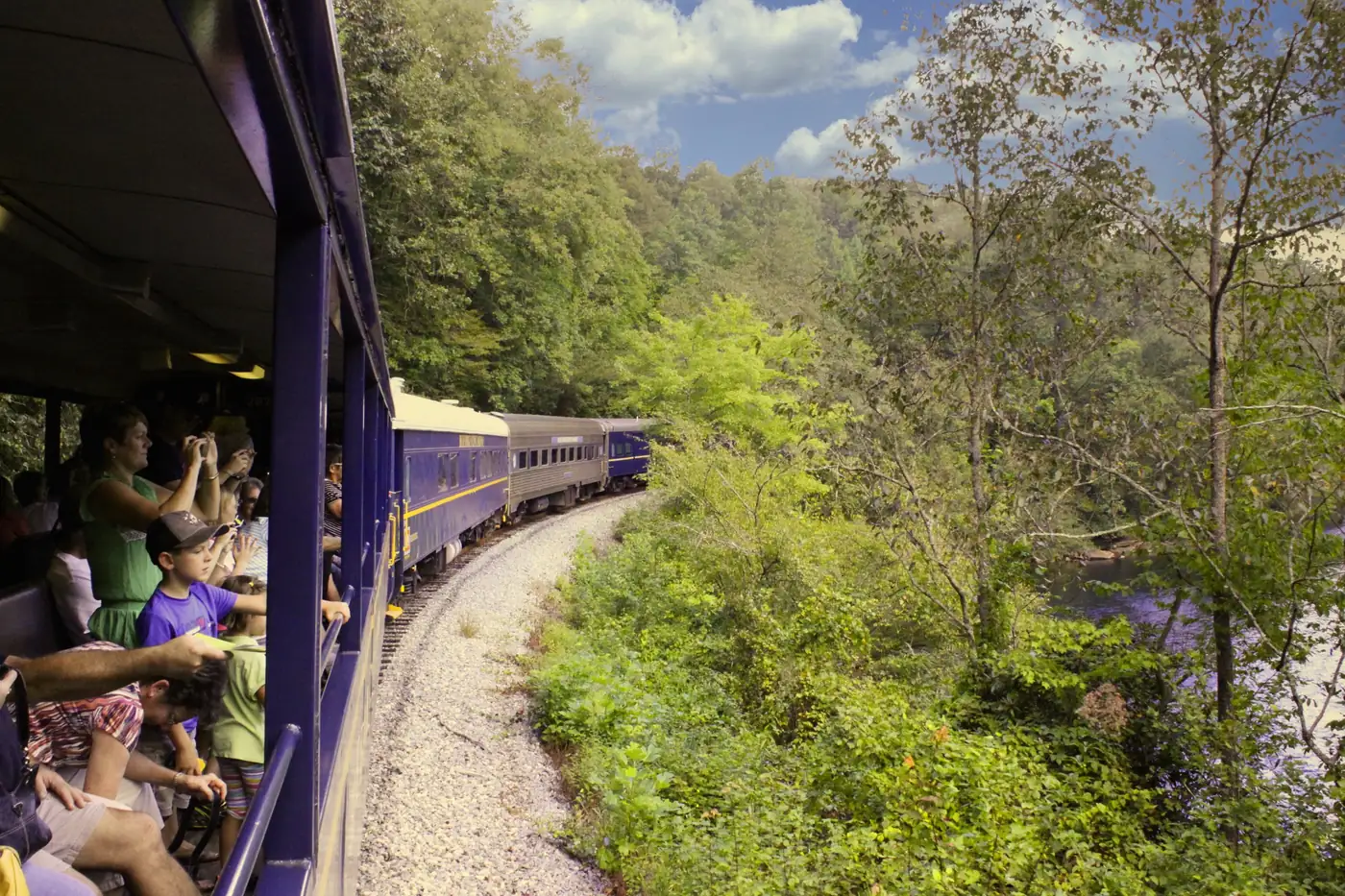Passengers in an open railcar look out as the train curves through green woods