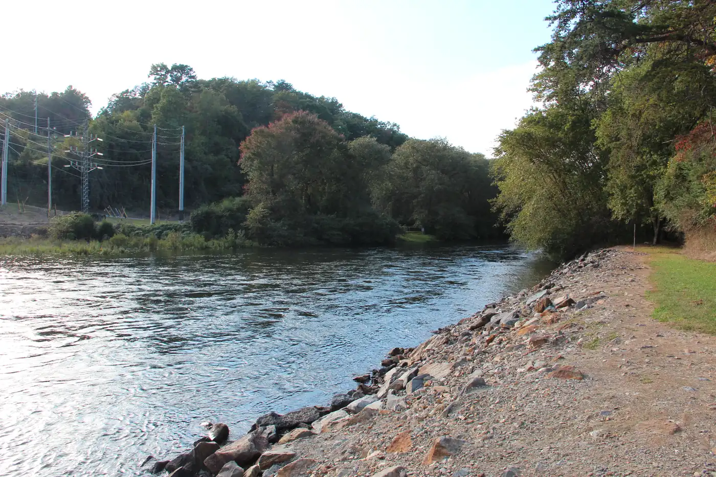 Clear river flowing past a gravel bank and leafy trees, with power lines on the far side