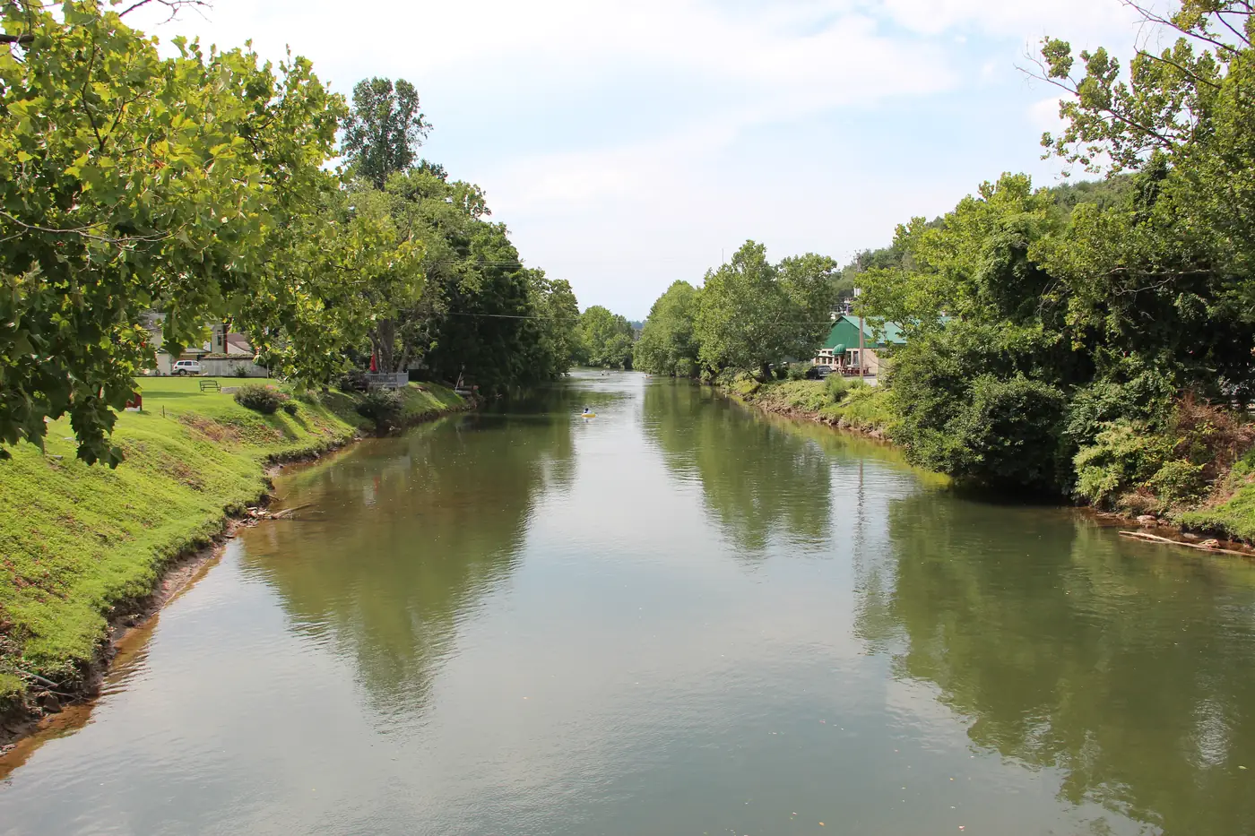 Calm green river between grassy banks and shade trees, with small-town buildings beyond