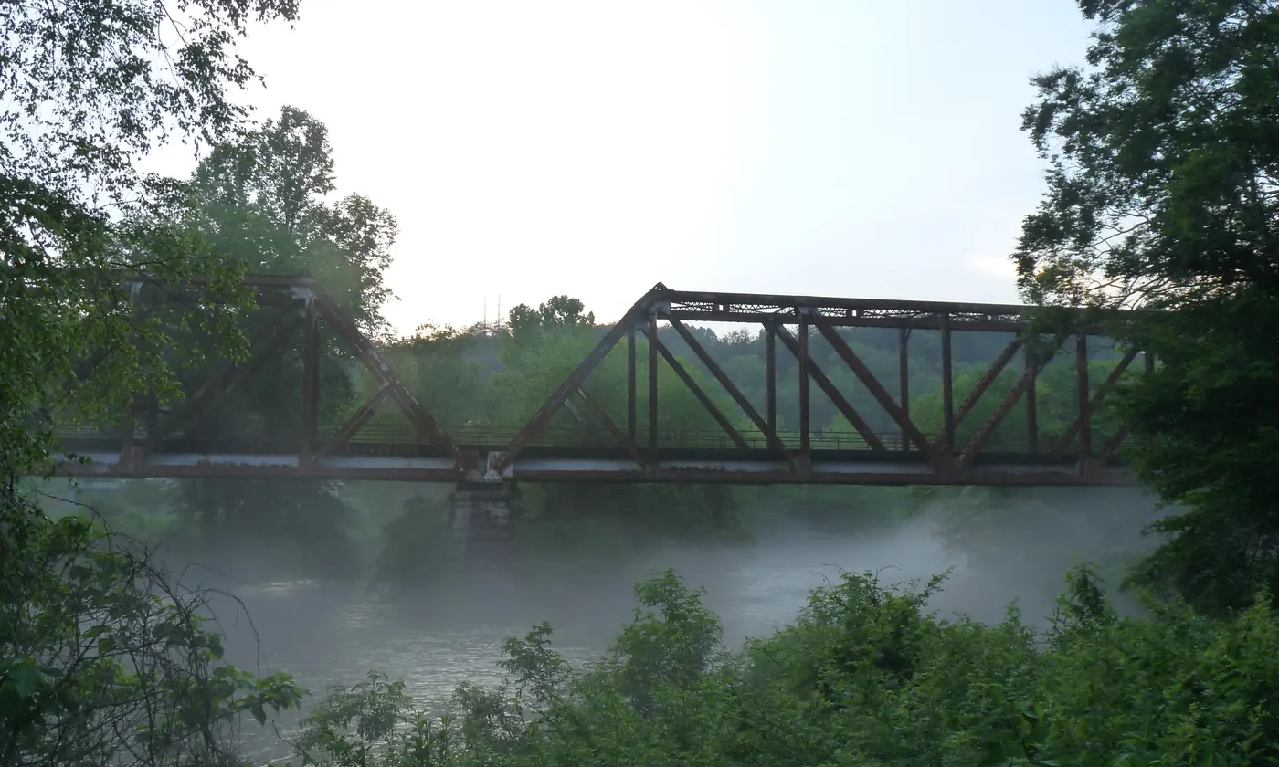 Steel truss railroad bridge over a misty river, framed by trees